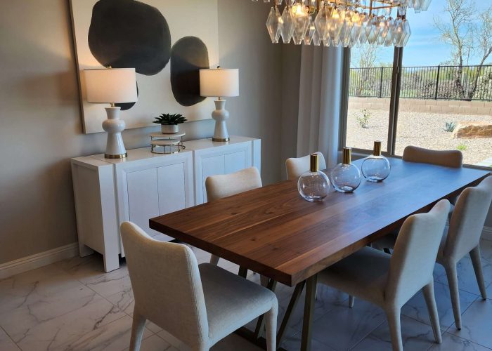 An elegant dining room with a dark brown rectangular table as the centerpiece, surrounded by bright white chairs. The table is adorned with a simple clear vase . Two white lamps sit on a white countertop against the wall, illuminating the space with a warm glow. Above the lamps hangs a modern abstract painting featuring three black dots on a white canvas. The room is bathed in natural light coming from a large window on the far wall, offering a serene view of the lush greenery in the yard outside.