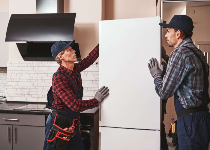 Two movers are shown in a kitchen, installing a refrigerator. One of the movers is holding the refrigerator in place, while the other is adjusting the height of the fridge feet. The fridge is free from any protective wrapping and its doors are closed. The kitchen cabinets and other appliances can be seen in the background.