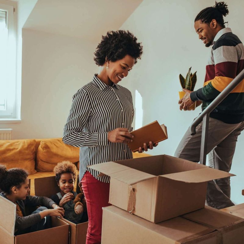 The image shows a family who has recently moved into a new home, unpacking boxes and organizing their belongings. The family consists of a couple and two young children. The adults are standing and removing items from a cardboard box, while the children are sitting in empty boxes in the background. The scene takes place in a residential setting, with other boxes and furniture visible in the background, indicating that the move is still in progress.